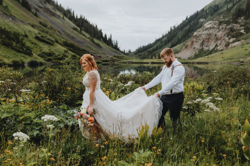 A groom holding his bride's dress as they walk through a field of flowers in Crested Butte, Colorado